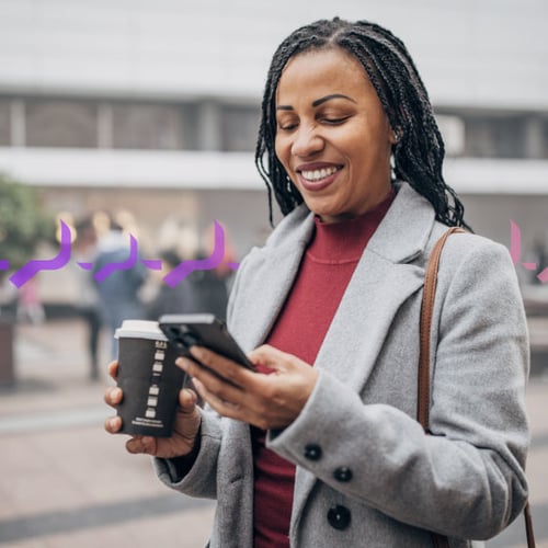 Traffic Control Blog Header Photo of a smiling woman outside of an office building, which is blurred in the background. She is looking at her phone in one hand and carrying a coffee cup in the other.  Across the frame in the background run a series of small purple icons representing text traffic.
